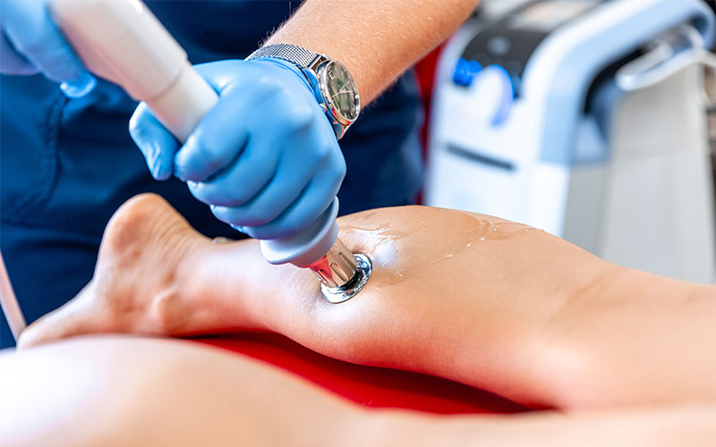 A healthcare professional, wearing blue gloves, applies a handheld device to a patient's leg, possibly for therapeutic treatment, in a medical setting with equipment visible in the background.