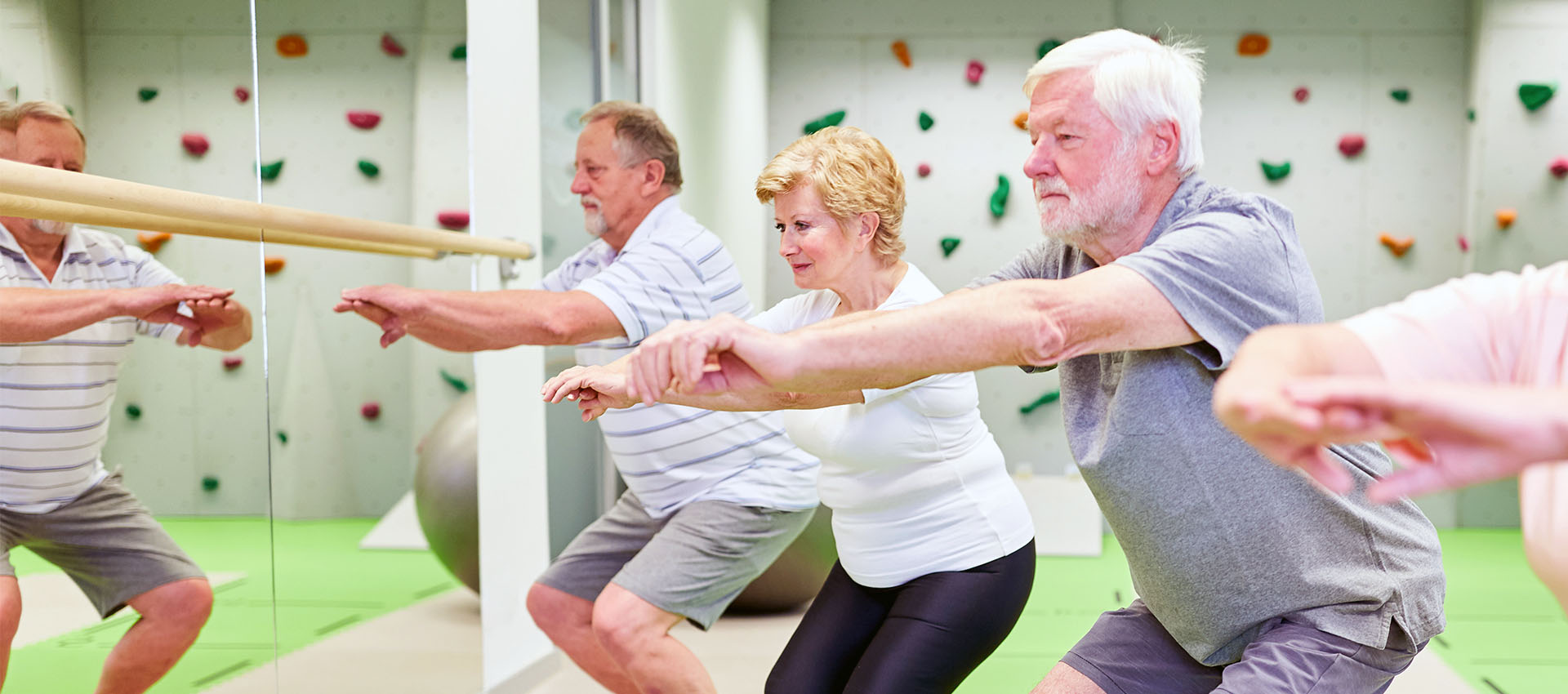 Three older adults exercise by doing squats in a brightly lit room with green flooring and climbing holds on the walls, facing a mirror and concentrating on their posture.