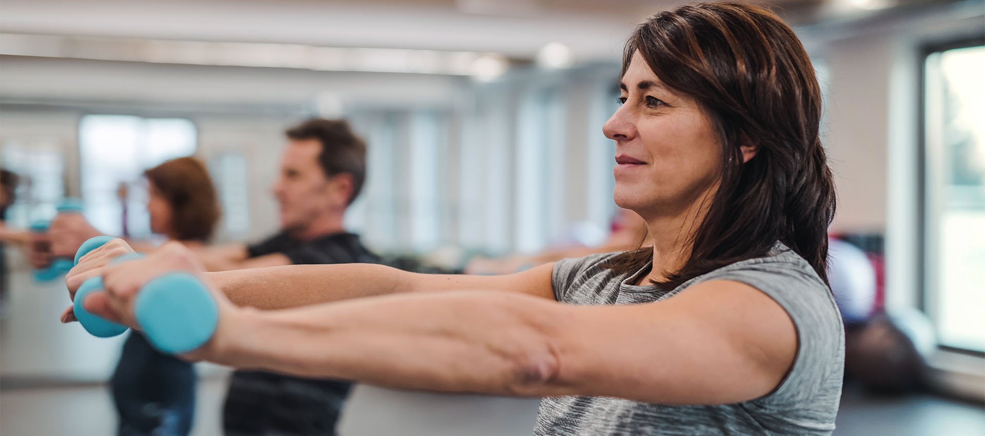 A woman lifts small blue dumbbells in a bright exercise room, with other people in the background similarly engaged in a fitness class.