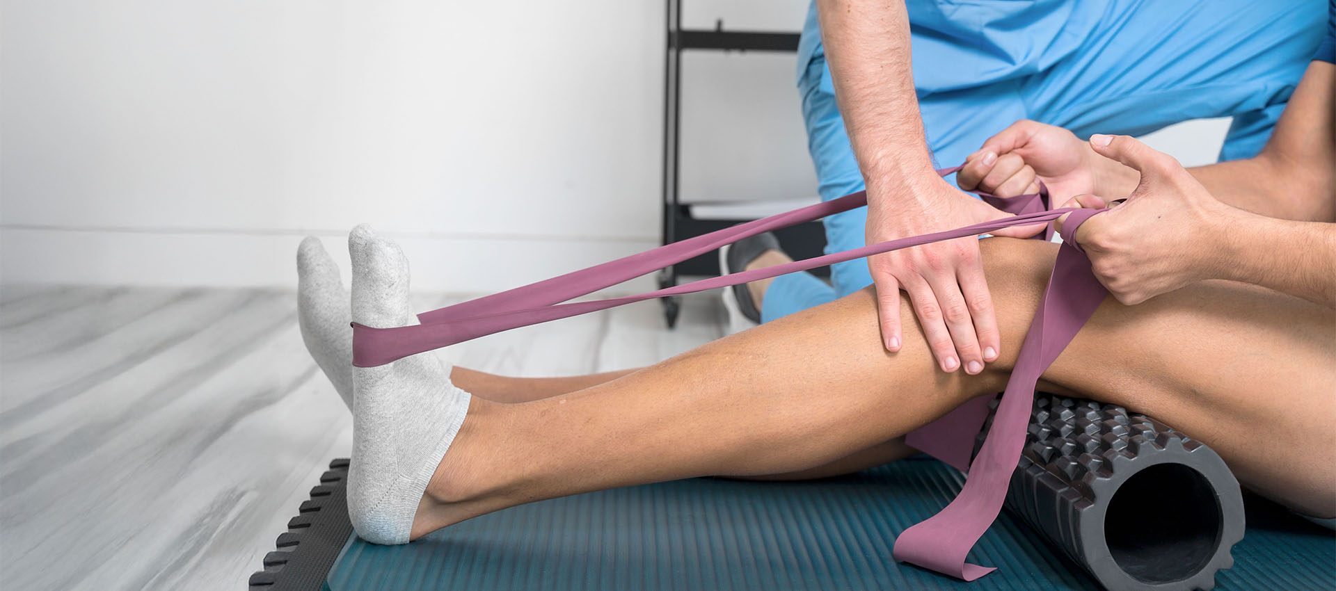 A person performs a leg stretch with resistance bands, assisted by a therapist, on a mat in a clinical setting. Nearby, a foam roller is placed under the calf.