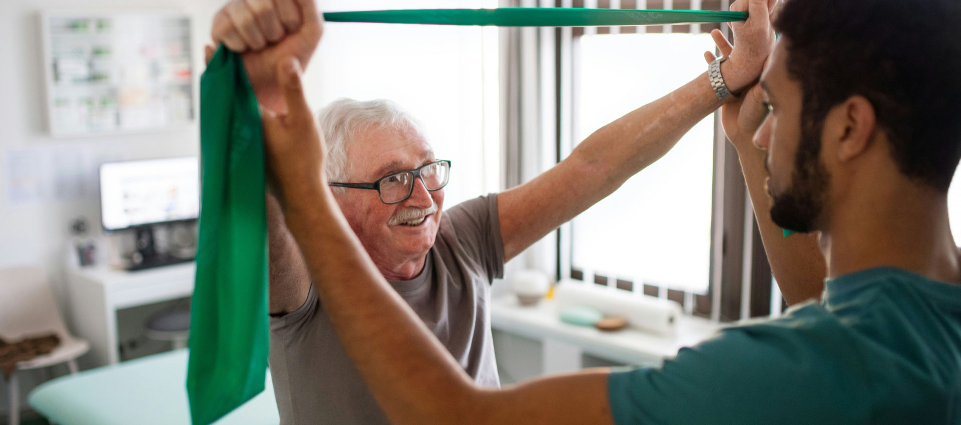 A man assists an elderly person in stretching a green resistance band, smiling in a bright therapy room with a desk and computer in the background.