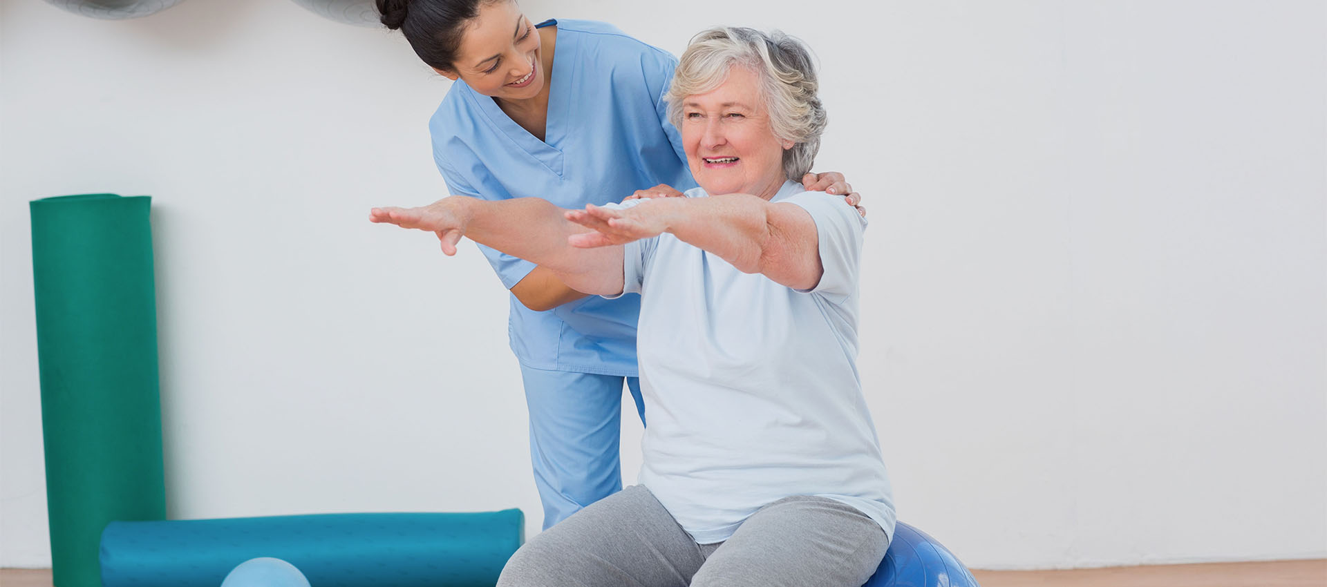 An older woman balances on an exercise ball with outstretched arms, assisted by a smiling healthcare professional in scrubs. Exercise mats and a white wall are in the background.