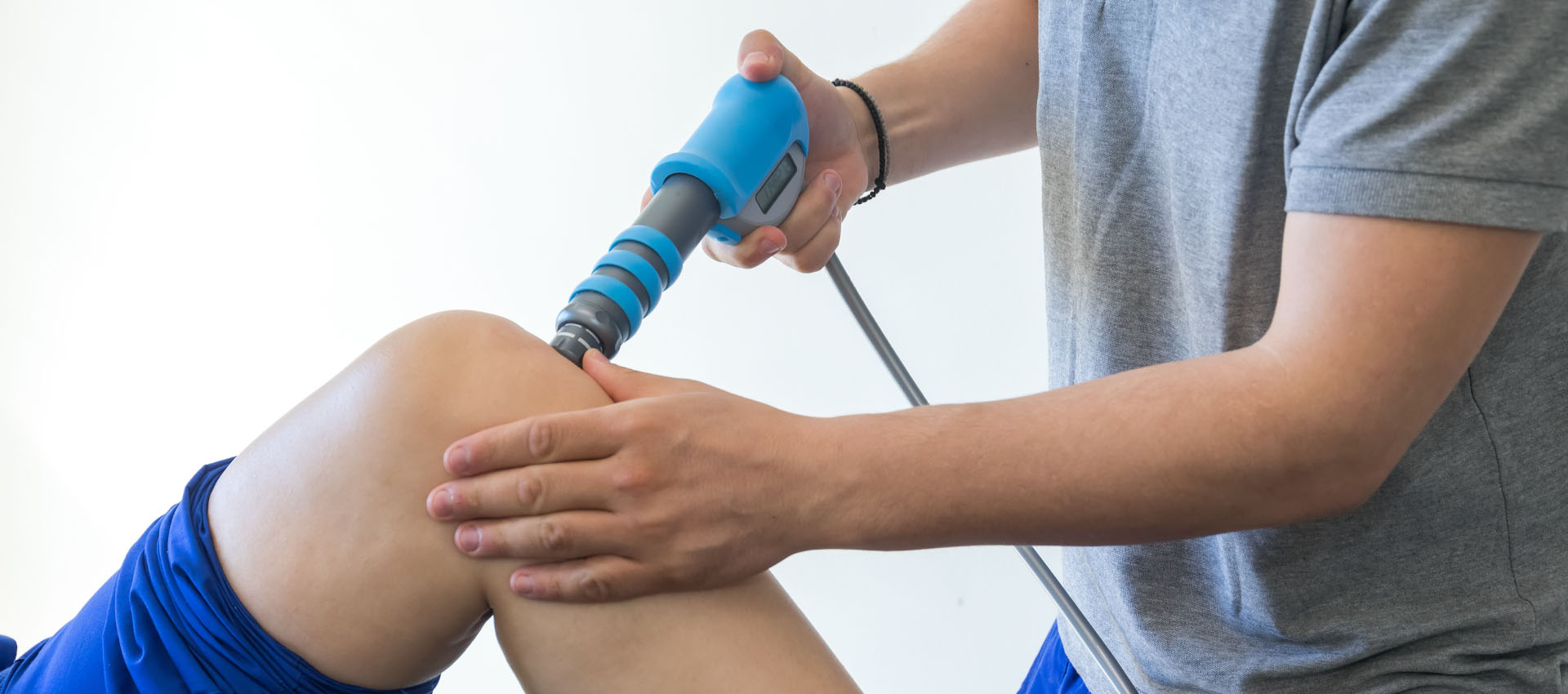 A healthcare professional applies a blue shockwave therapy device to a patient's knee, ensuring proper contact. The patient is seated, wearing blue shorts, in a clinical environment.