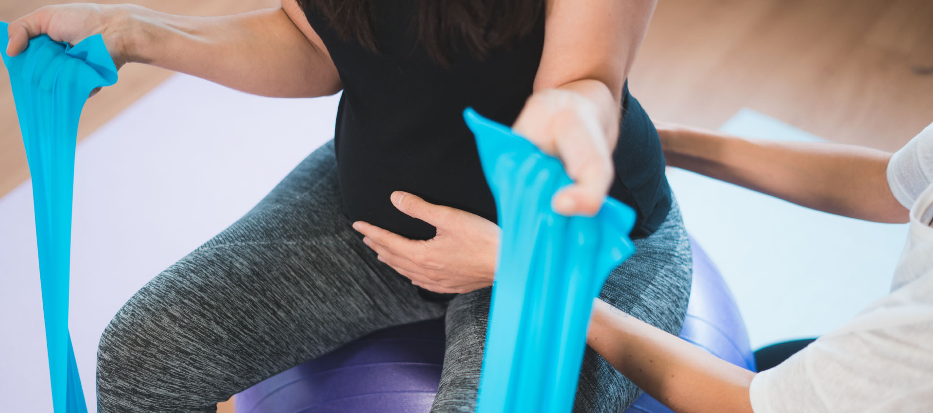 A person sits on an exercise ball, pulling a blue resistance band, assisted by another person in a light-filled room.