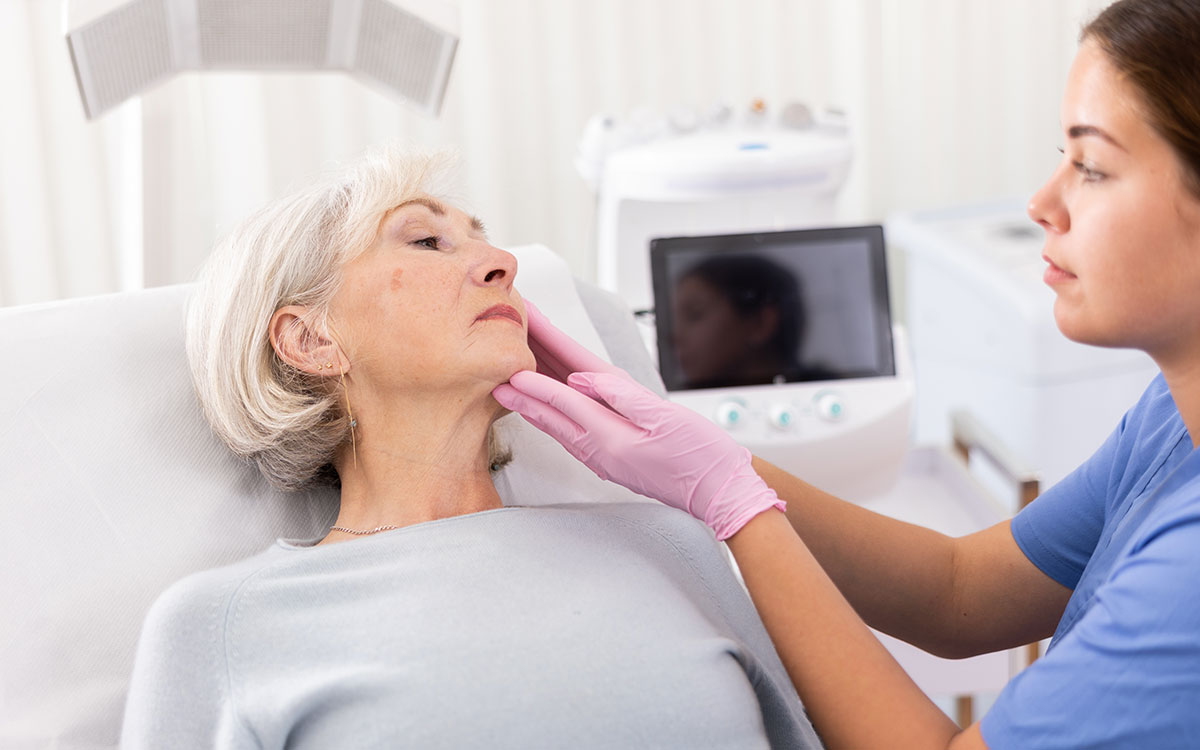 A healthcare professional in blue scrubs gently examines an elderly woman's jawline, wearing pink gloves. They are in a clinical setting with medical equipment visible in the background.
