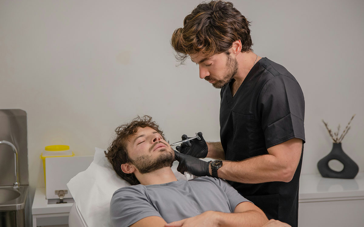 A healthcare professional examines a patient's throat in a clinical setting, wearing black gloves. The patient is lying on an examination table with white bedding, indicating a medical environment.