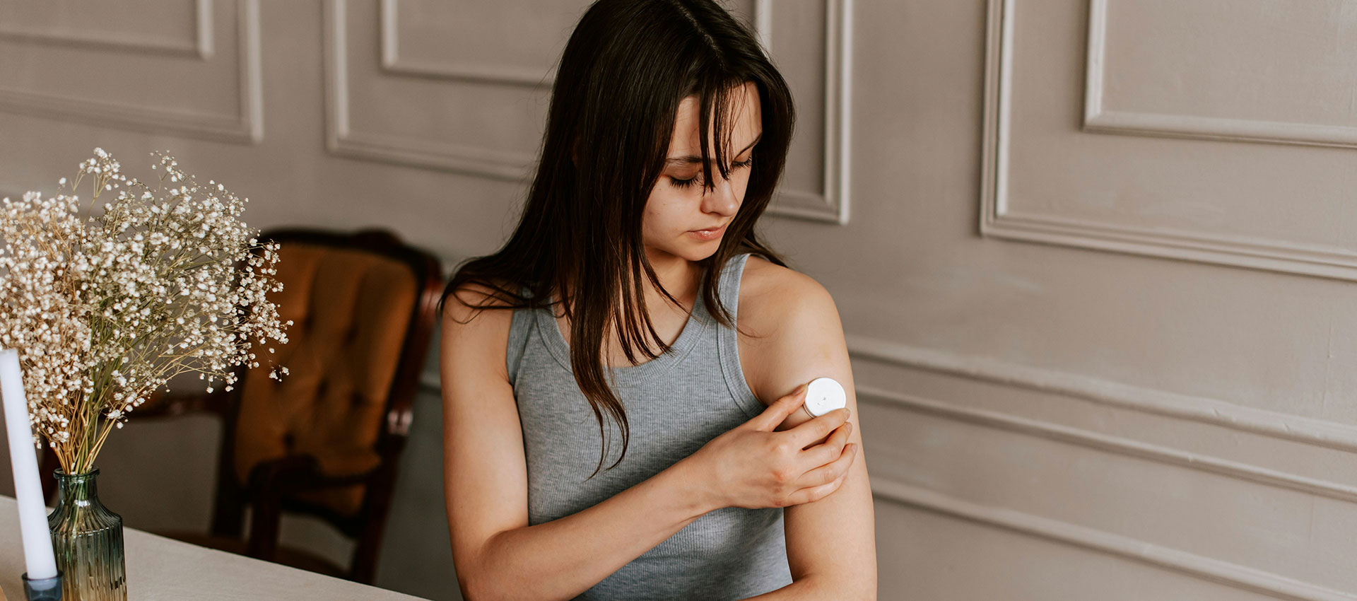 A woman applies lotion to her arm while seated at a table. A vase of flowers and an ornate chair are visible in the background, set against a paneled wall.