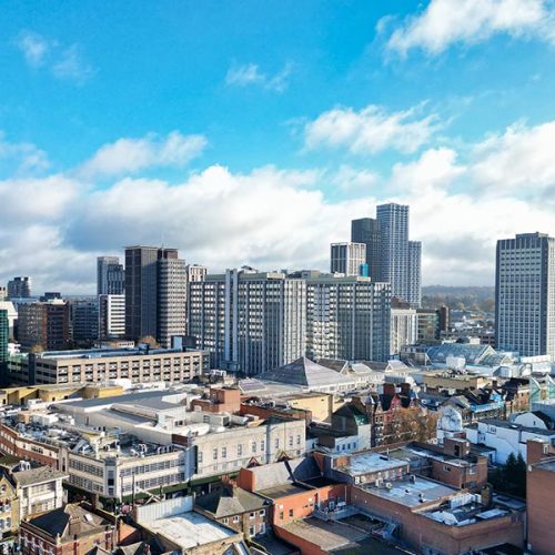 A cityscape with tall modern buildings under a bright blue sky dotted with clouds, surrounded by smaller structures, suggesting a vibrant urban environment.