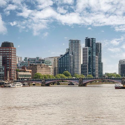 A modern city skyline with tall buildings stands against a partly cloudy sky, overlooking a wide river crossed by a bridge.