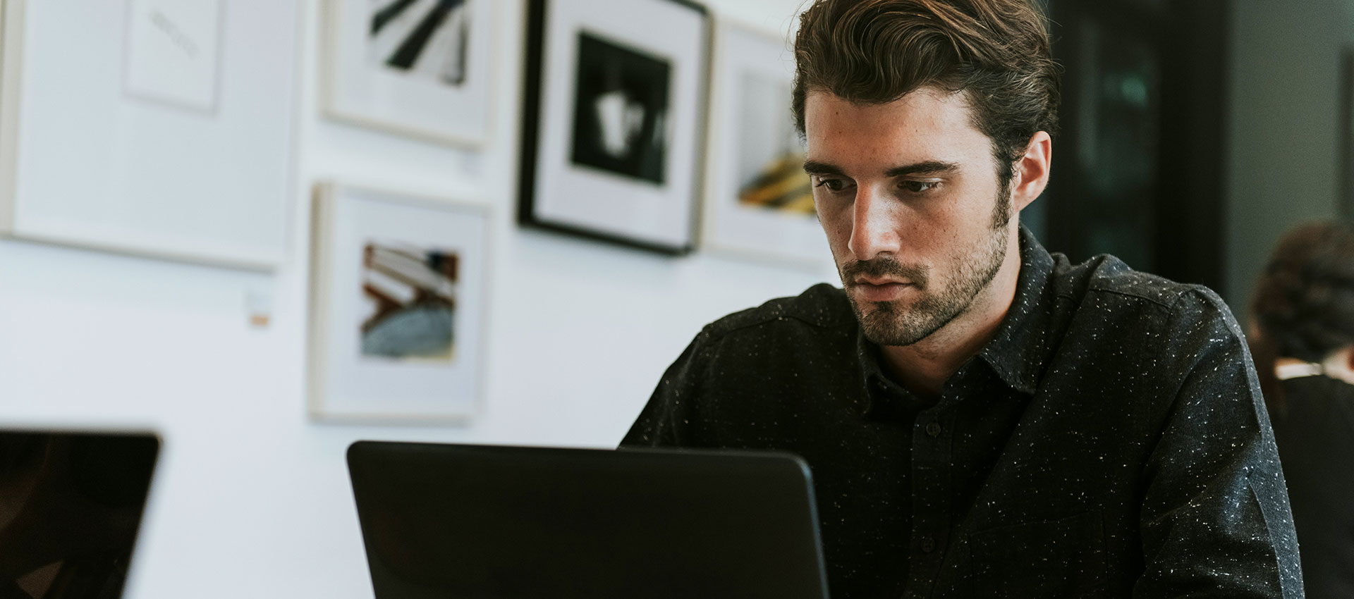 A person sits focused on a laptop in an art-filled room, surrounded by framed pictures on the wall, creating a modern and creative workspace atmosphere.