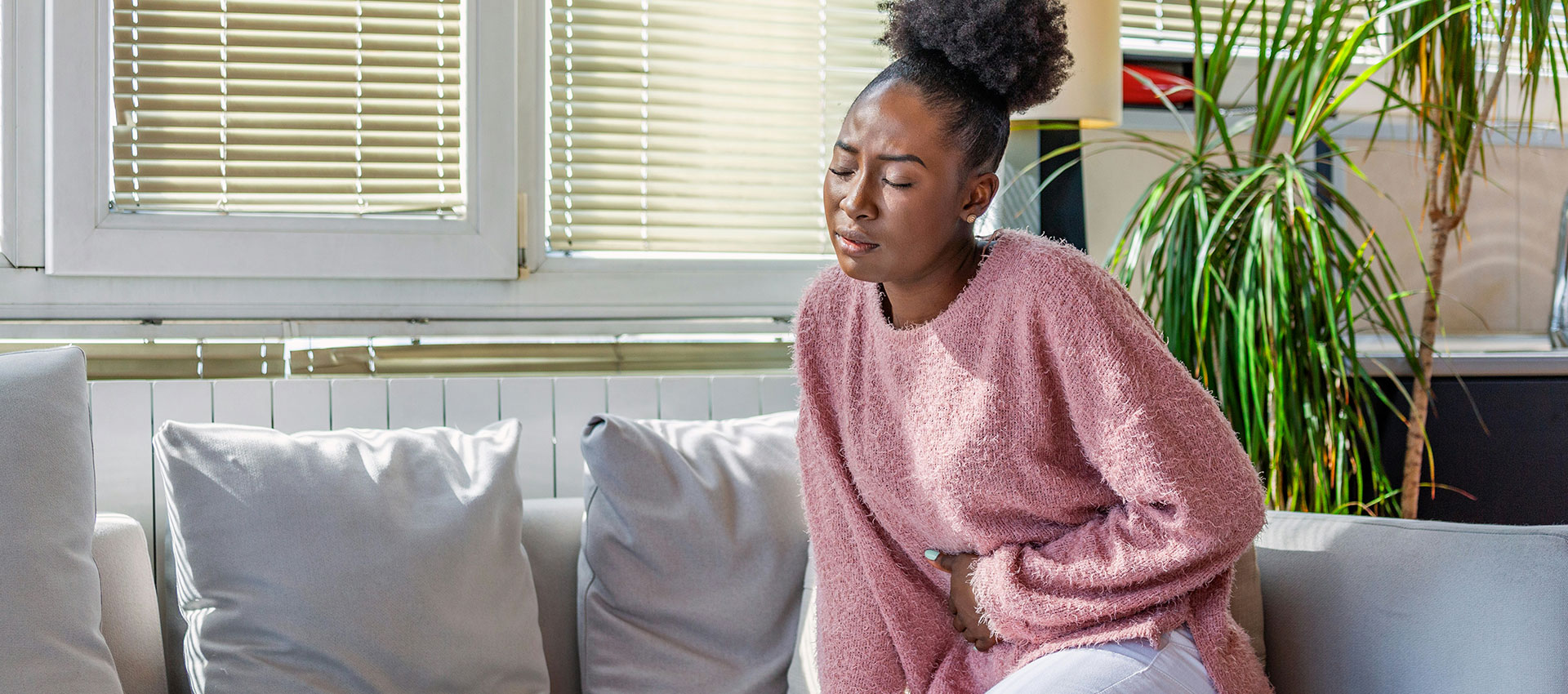 Woman in a pink sweater sits on a couch, wincing and clutching her side, suggesting discomfort. The room has large windows with blinds and potted plants nearby.