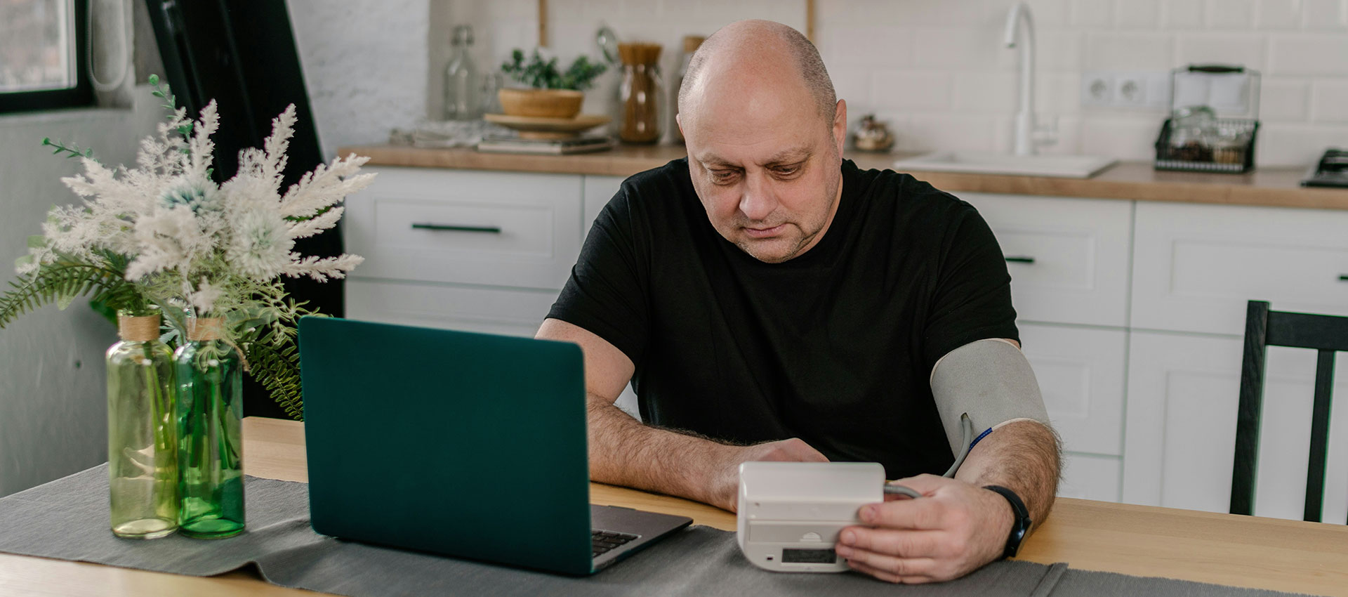A man checks his blood pressure using a monitor at a kitchen table, with a laptop nearby and decorative plants in the background.