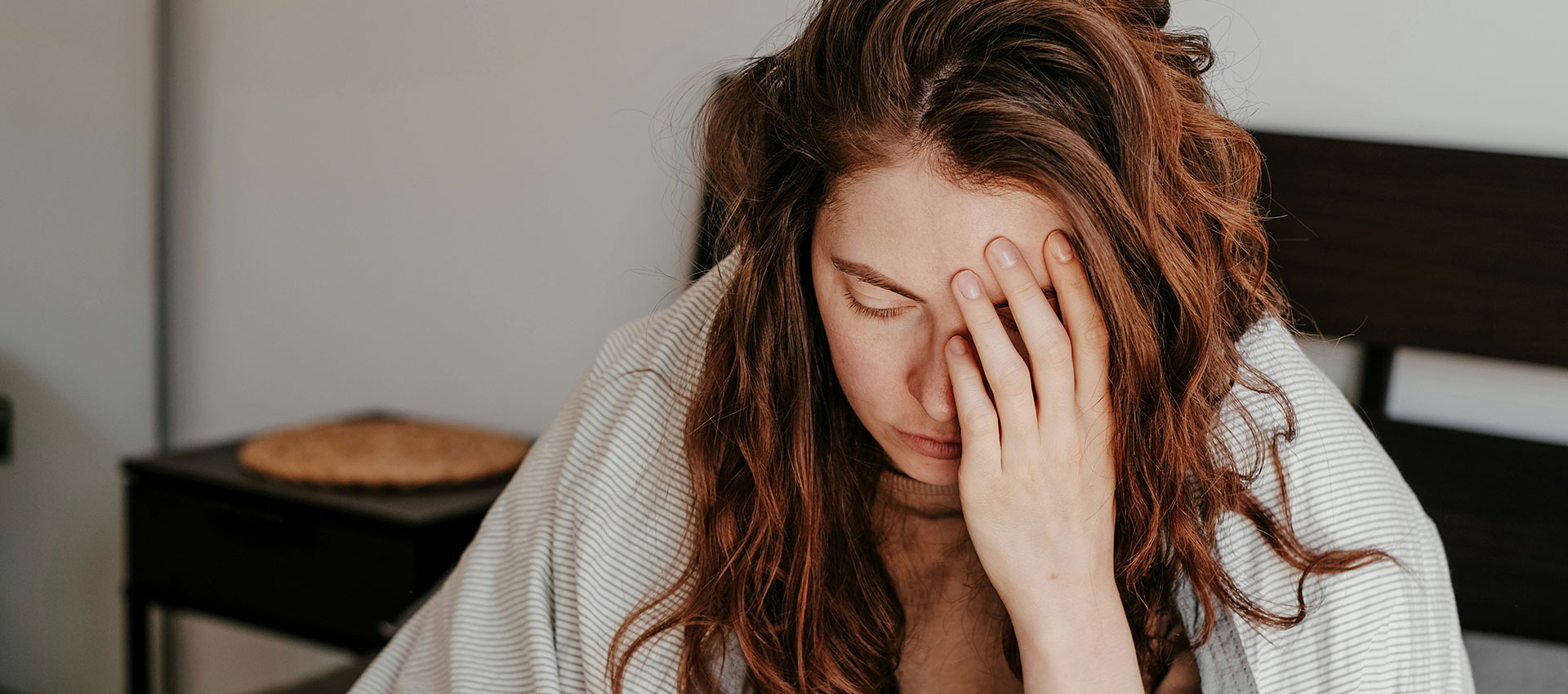 A person sits on a bed with their hand on their face, appearing tired or stressed, in a bedroom with a nightstand in the background.
