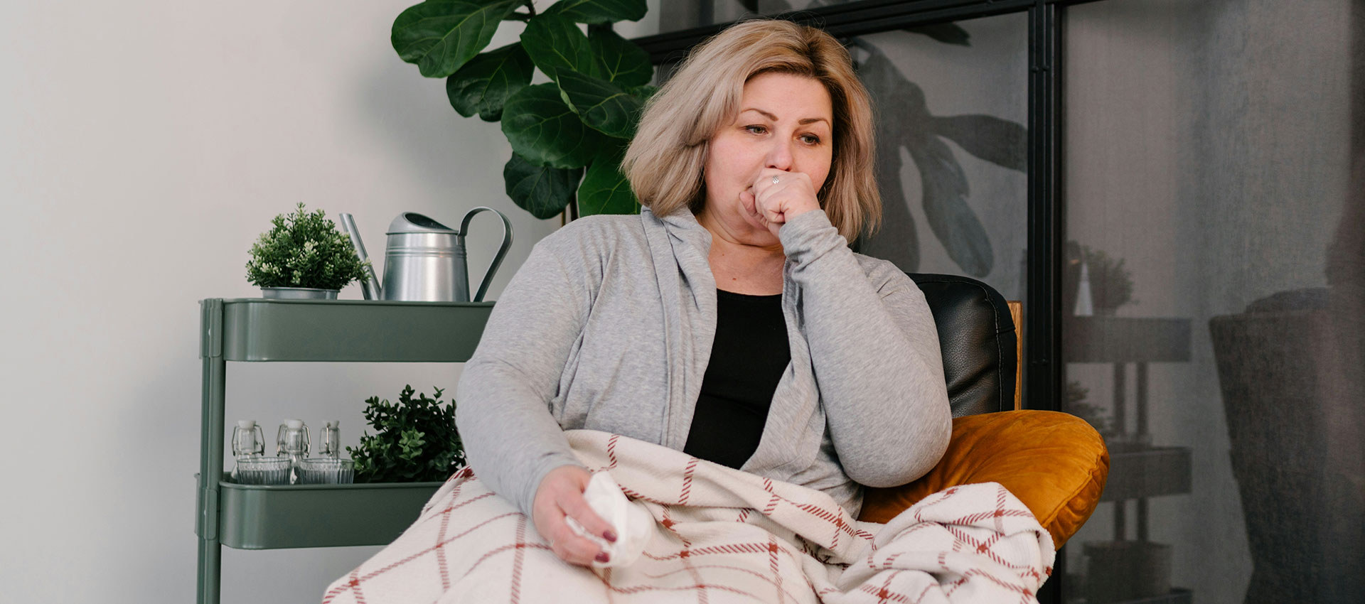 A woman, sitting on a chair, coughs into her hand while wrapped in a plaid blanket. Nearby, a green cart holds plants and a watering can, with a leafy plant in the background.
