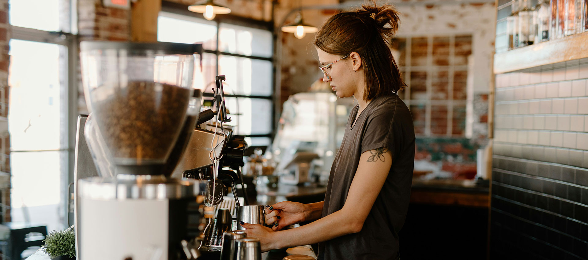 A barista prepares coffee using a machine in a rustic cafe with large windows and exposed brick walls.