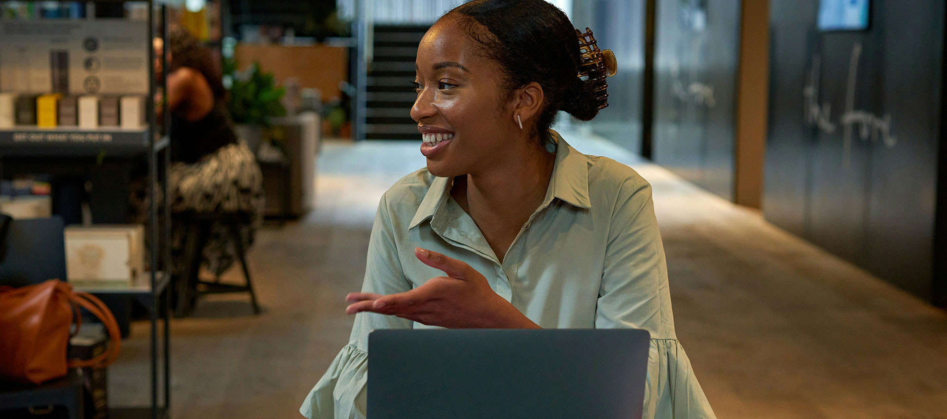 A person sits at a table with an open laptop, gesturing while talking with another unseen individual. The setting is a modern indoor environment with soft lighting and plants.