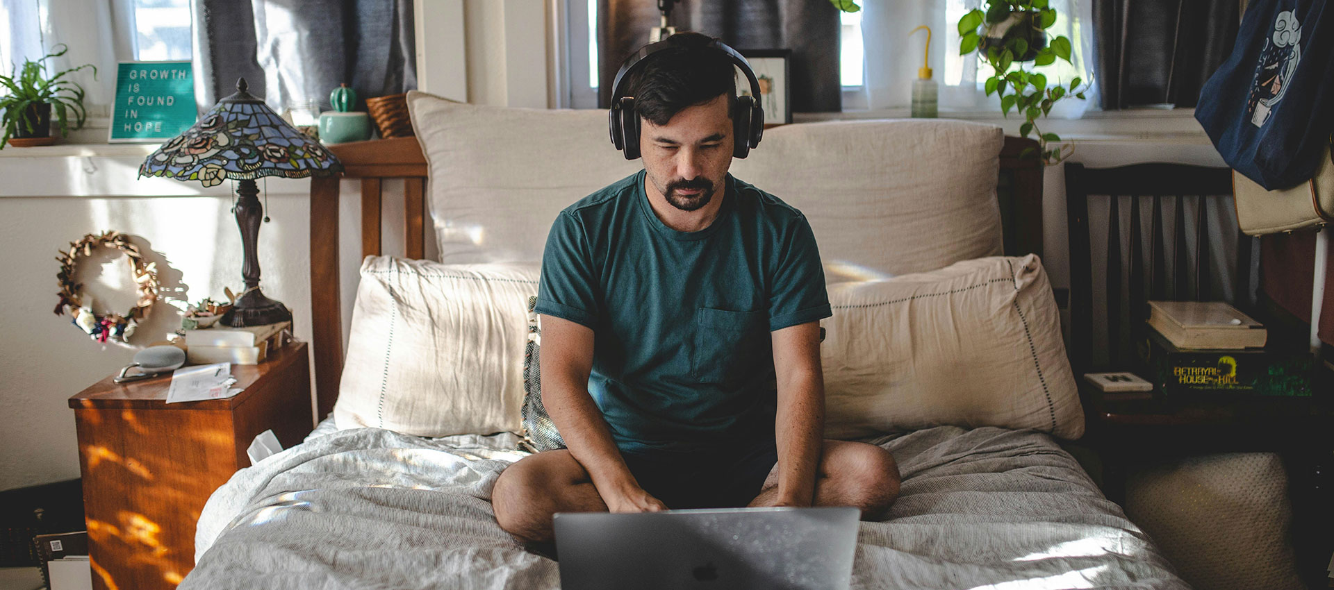 A person sits cross-legged on a bed in a cozy room, concentrating on a tablet. Soft light filters through a nearby window, creating a peaceful atmosphere.