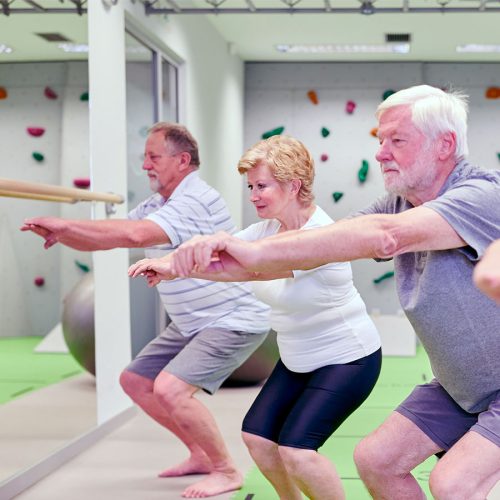 Three older adults exercise by doing squats in a brightly lit room with green flooring and climbing holds on the walls, facing a mirror and concentrating on their posture.