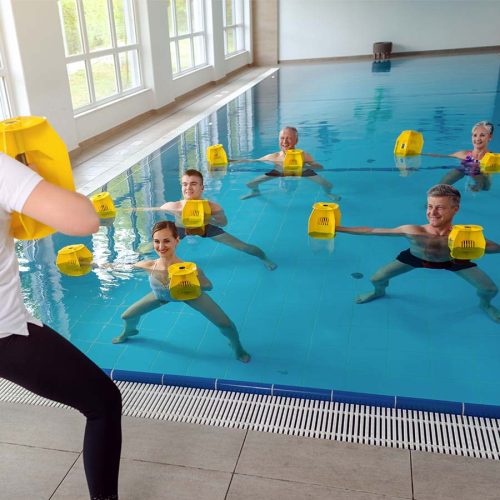 People exercise in a swimming pool, each holding yellow foam weights. A person leads the session from the poolside. Large windows line the wall, providing natural light.