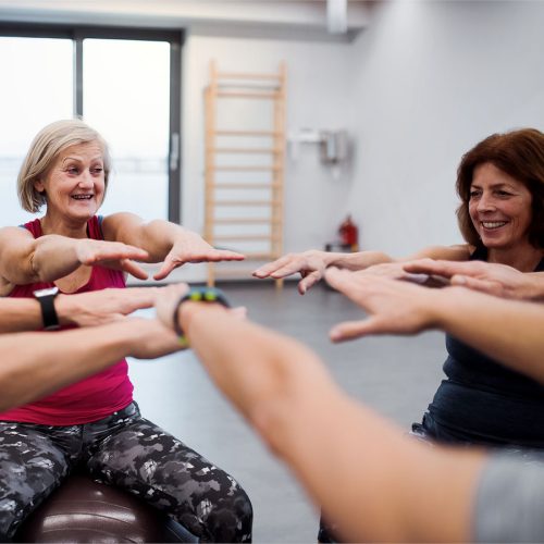 A group of individuals sits in a circle, smiling and extending their arms toward each other in a bright gym setting. Fitness equipment is visible in the background.