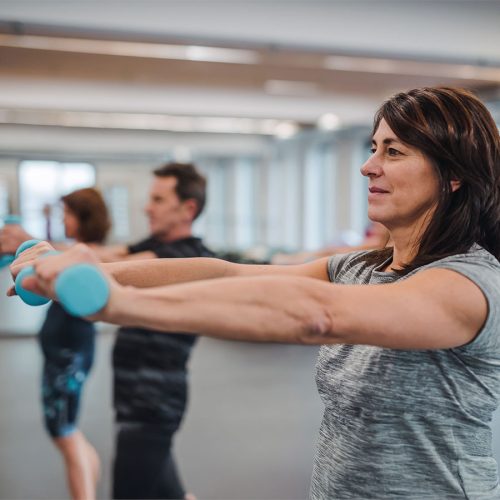 A person lifts light blue dumbbells in an exercise class, focusing on strength training. Others in the background participate, creating a group fitness setting in a bright, spacious room.