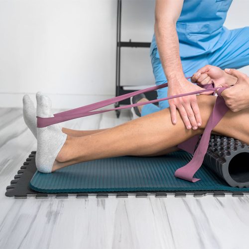 A person assists another in stretching using a resistance band around their foot on a mat, in a clinical setting with light wood flooring.