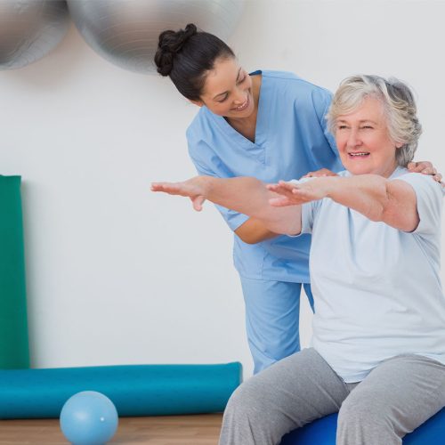 A healthcare professional assists an elderly woman performing arm exercises on a stability ball in a bright room with yoga mats and a small exercise ball in the background.