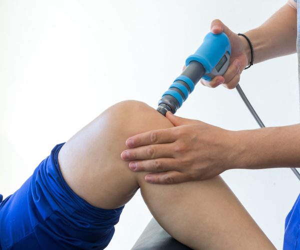 A hand holds a blue therapy device, applying treatment to a person's knee in a clinical setting. The patient is wearing blue shorts, lying back on an examination table.