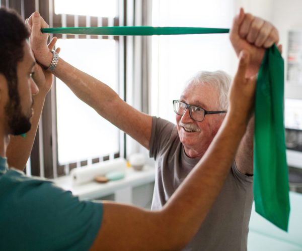 An elderly man exercises with a green resistance band, assisted by a healthcare professional, in a bright room with large windows.