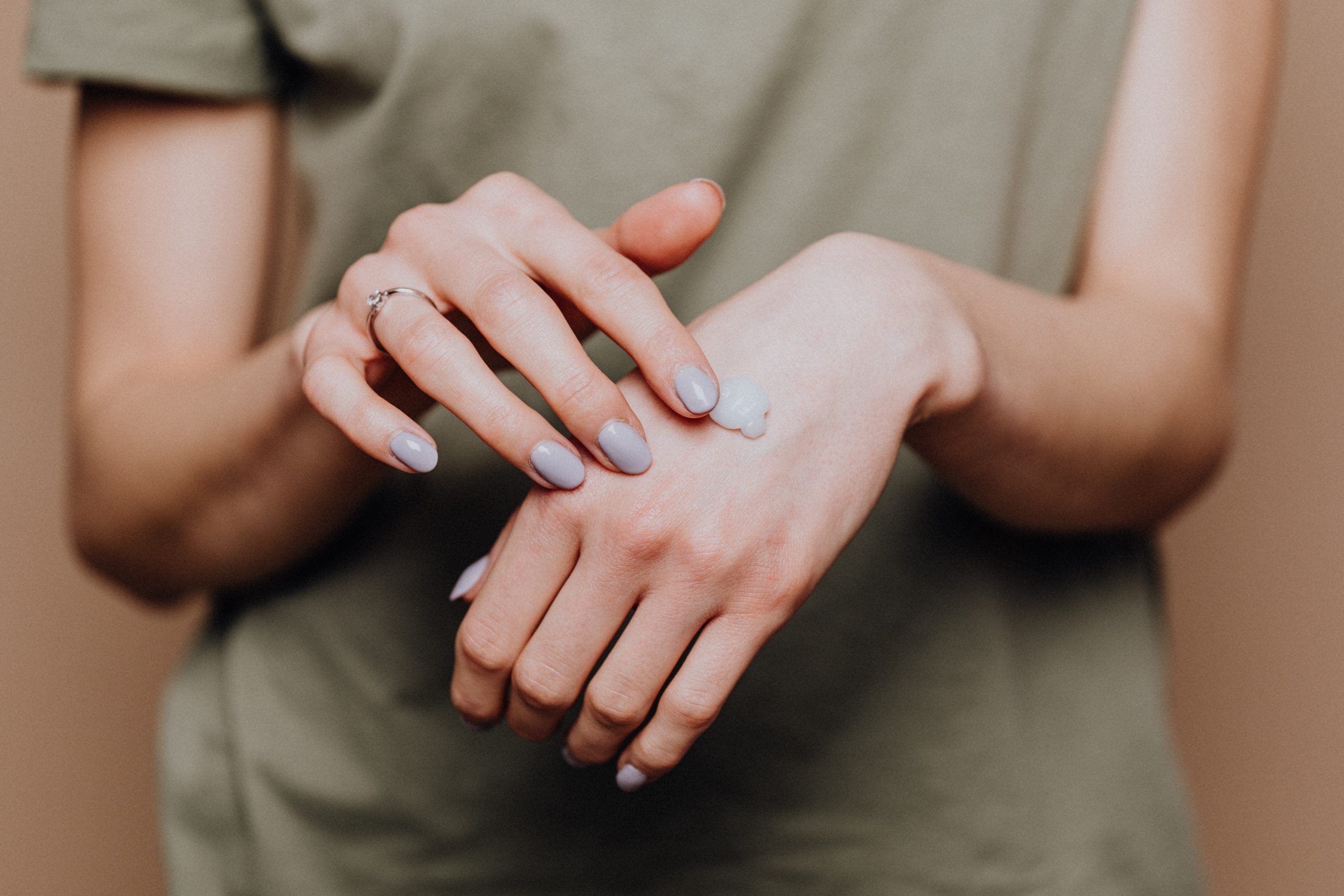 Hands applying cream on the back of one hand in a gentle motion, with a beige background. The person wears a green shirt and a ring on one finger.