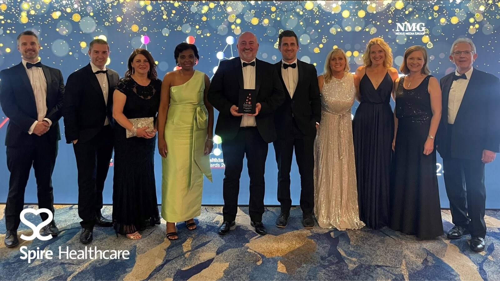 A group of ten people in formal attire pose together; one person holds an award. They're in front of a backdrop with confetti-like lights and logos for Spire Healthcare and NMG.