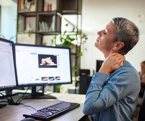 A person with short hair massages their neck while sitting at a desk with two computer monitors, surrounded by office plants and shelves.