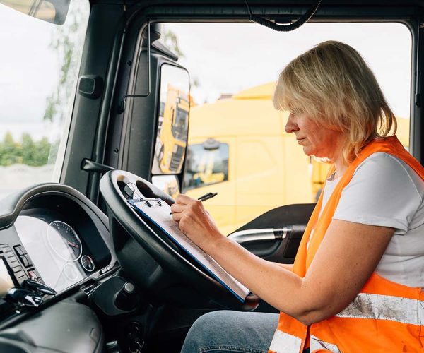 A person in an orange safety vest writes on a clipboard inside a truck. The vehicle dashboard and window view of another truck are visible.