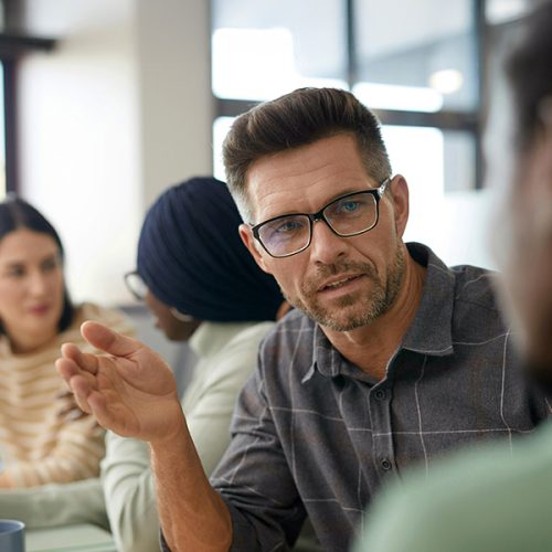 A man wearing glasses gestures while speaking to a person in a professional meeting. Two other people are in the background, seated at a table in a bright office.