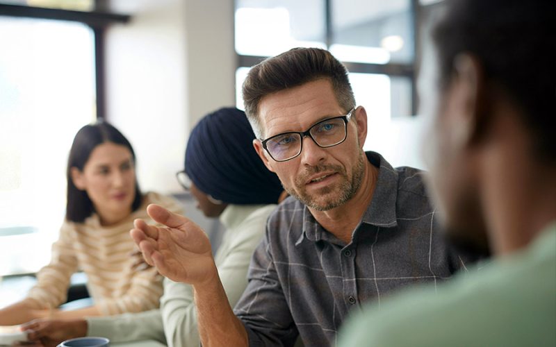 A man wearing glasses gestures while speaking to a person in a professional meeting. Two other people are in the background, seated at a table in a bright office.
