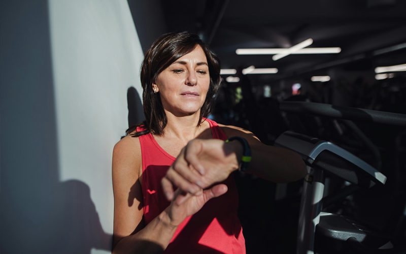 Woman in a red tank top checks a smartwatch while standing beside exercise equipment in a dimly lit gym.