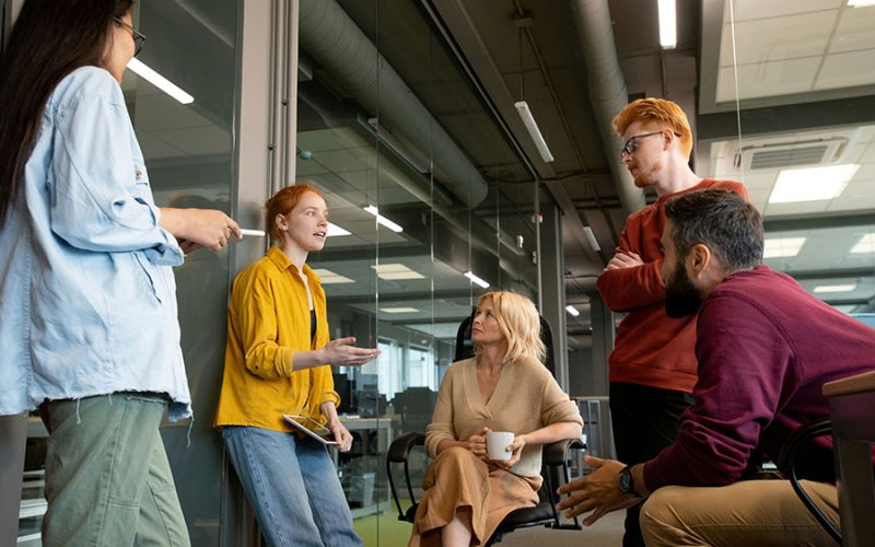 A group of four colleagues engage in discussion inside a modern office, with glass walls and overhead lighting. One person stands holding a notebook, while the others are seated, listening attentively.