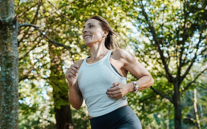 A woman jogs through a forested area, wearing a white tank top and dark leggings, surrounded by trees with green leaves, under a sunny sky.