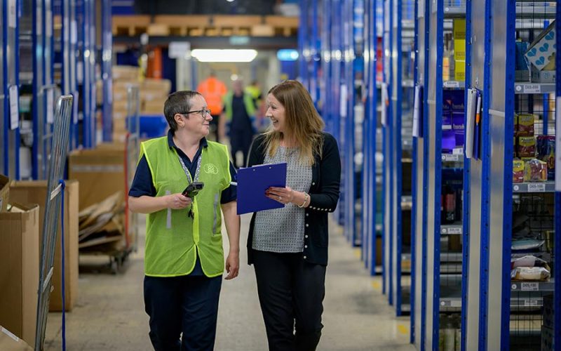 Two people walk and converse in a warehouse aisle. One holds a clipboard, and the other has a handheld scanner. Shelves with various items line the walkway.