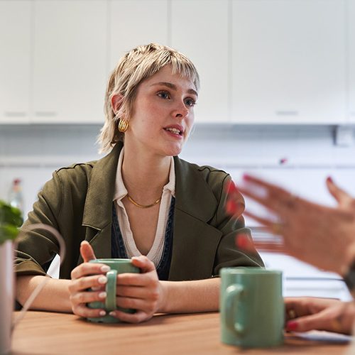 A person holding a green mug is sitting at a table, engaged in conversation with someone gesturing, in a kitchen setting with white cabinets.