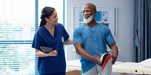 A healthcare professional in blue scrubs, holding a tablet, assists a man performing a leg stretch in a brightly lit room with medical equipment and large windows.