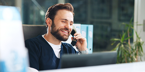 A man is sitting at a desk, smiling while talking on a phone. He is in an office environment with binders and a plant visible in the background.