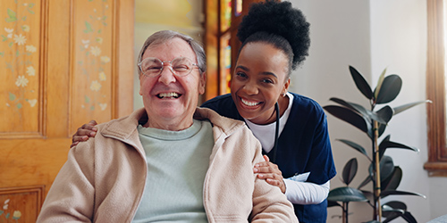 A caregiver in a blue uniform smiles warmly while standing behind an elderly man seated and laughing. They are indoors, with a wooden door and houseplant in the background.