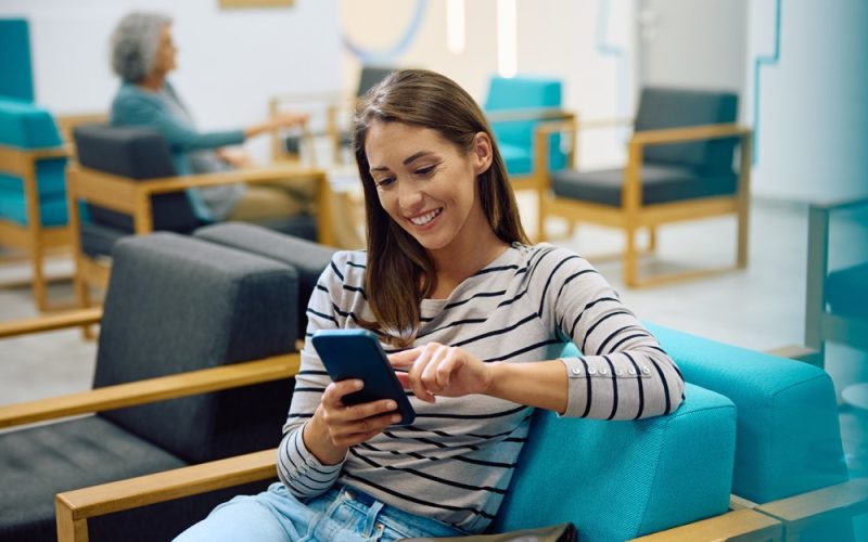 A woman is sitting on a modern chair using a smartphone, smiling. In the background, an older person is seated, creating a relaxed waiting room atmosphere.