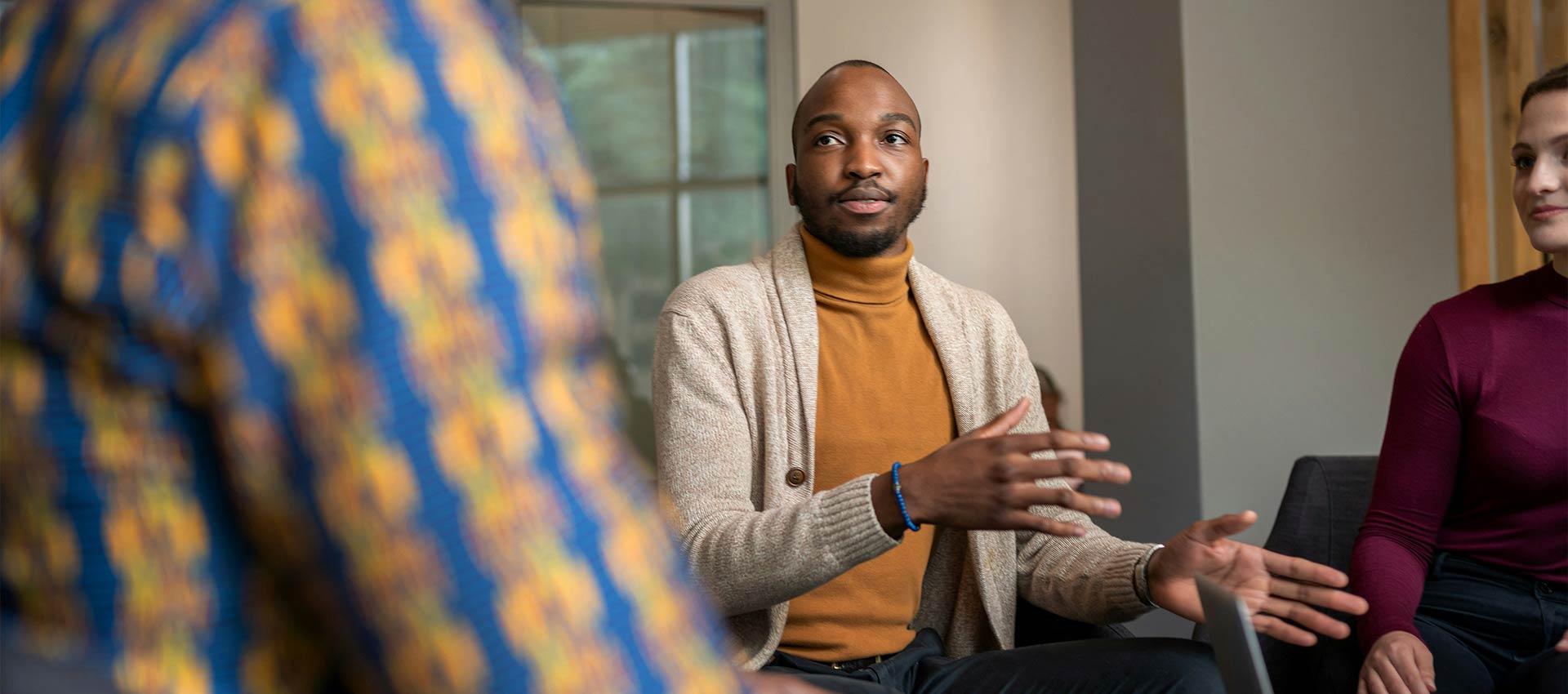 A person gestures while speaking in a group setting; two others listen attentively, seated nearby in a cozy room with neutral-colored walls. No text is present.