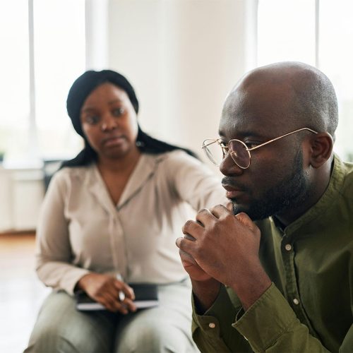 A man in glasses sits thoughtfully with hands clasped, while a woman beside him offers support, placing a hand on his shoulder in a bright room.