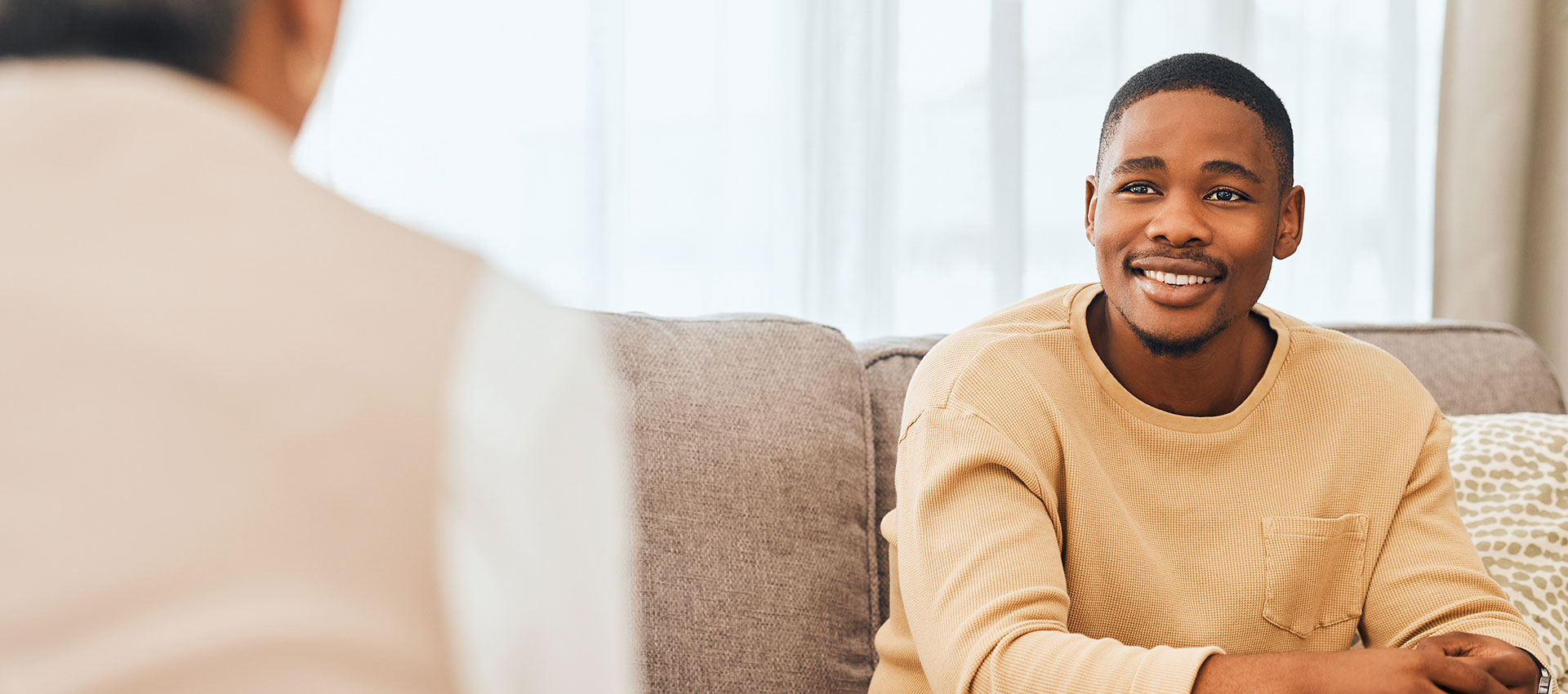 A man in a tan sweater smiles while sitting on a couch, speaking to another person in an indoor setting with soft lighting.