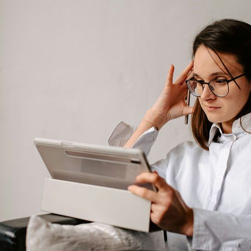 A person wearing glasses is seated, holding a tablet and resting their head on their hand, appearing focused. The background is a plain, light-colored wall.
