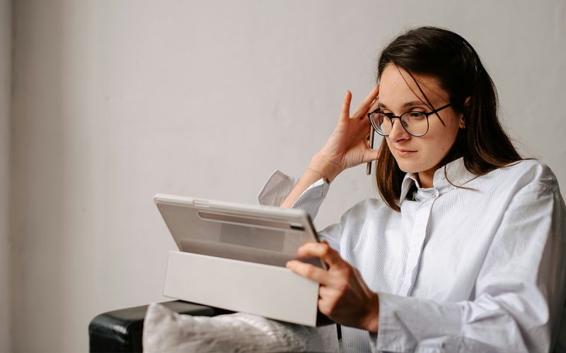 A person wearing glasses is seated, holding a tablet and resting their head on their hand, appearing focused. The background is a plain, light-colored wall.