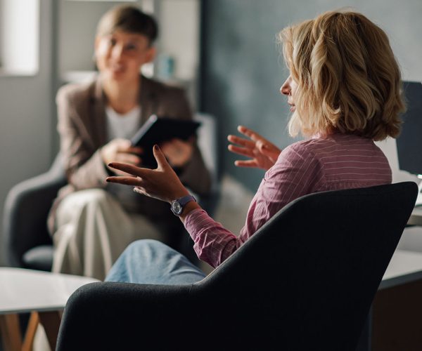 Two people sit in armchairs, engaged in a conversation. One person gestures with their hands while the other holds a tablet. The setting appears to be an office or therapy room.